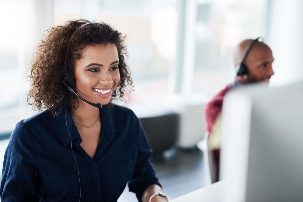 Smiling female call center agent wearing a headset, working at a computer in a bright office.