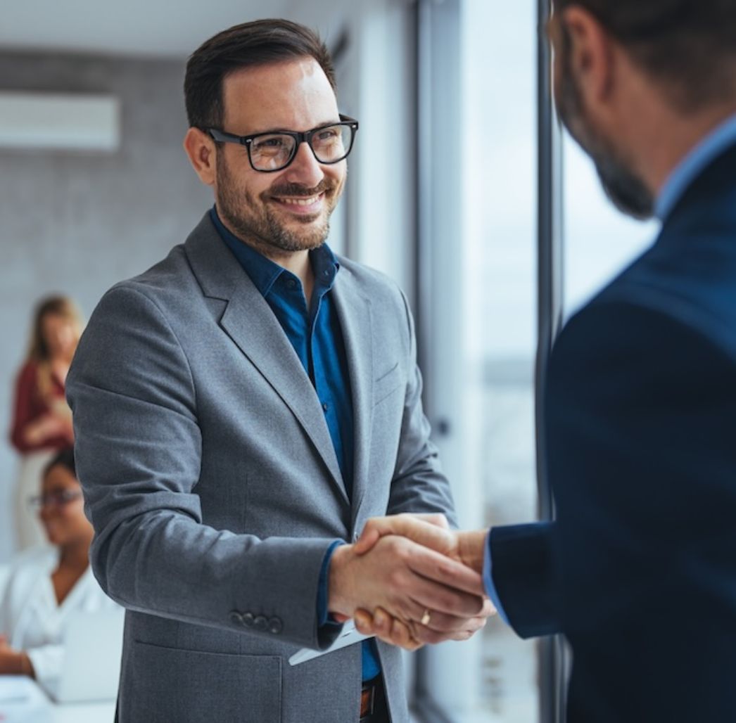 Smiling businessman in a grey suit shaking hands with a colleague in an office setting, with coworkers working in the background.