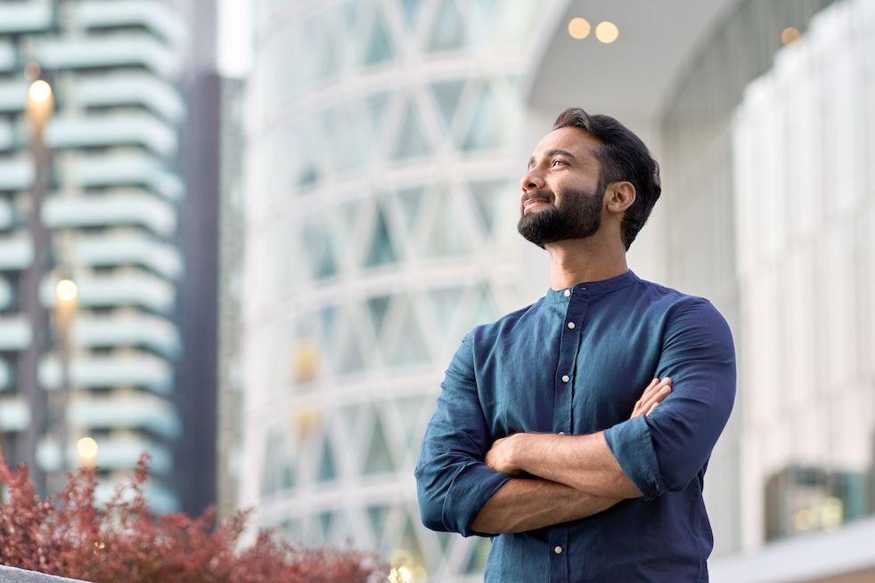 Confident man standing outdoors in a city with arms crossed, looking upward with a thoughtful expression.