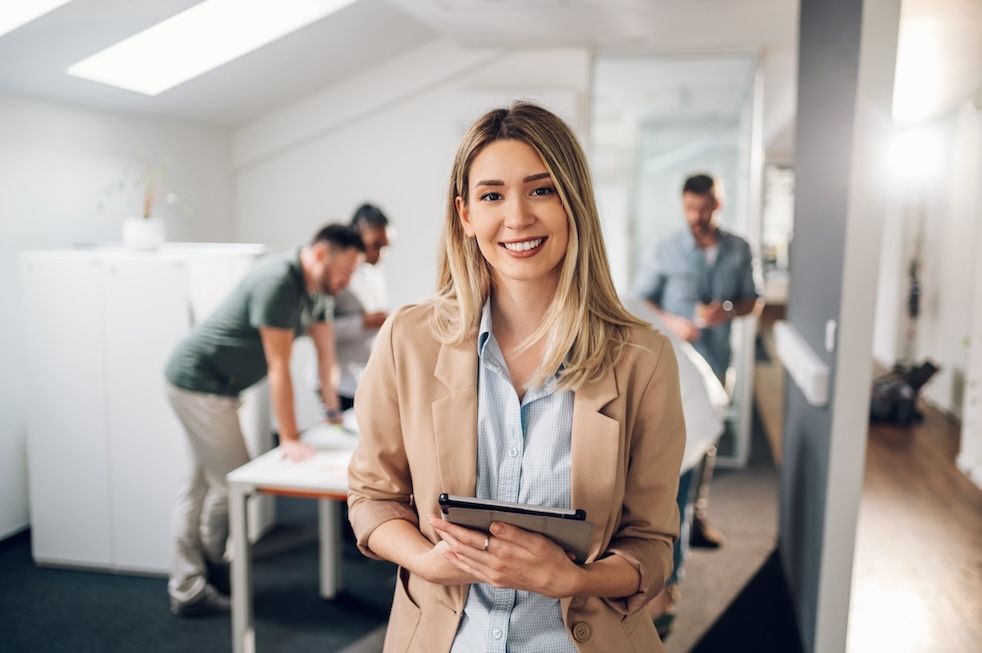 Smiling young woman holding a tablet in a modern office, with three colleagues working in the background.